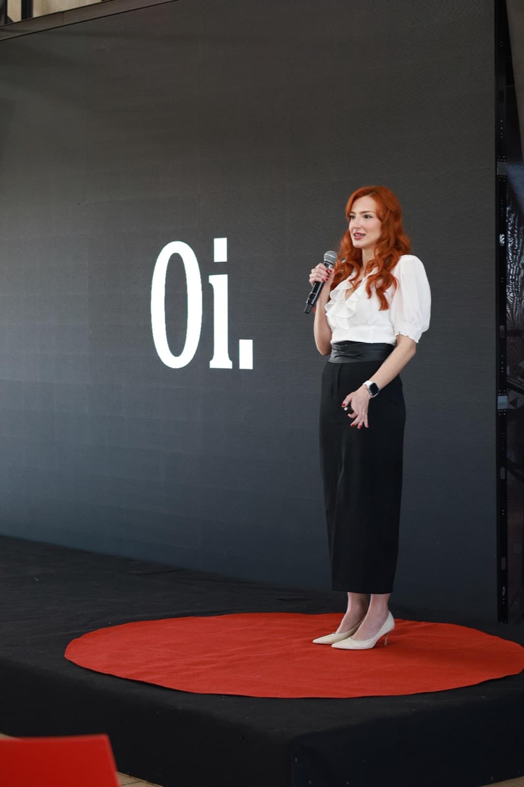 Eu no palco da TEDxUFTM, no tapete vermelho, com o slide “Oi.” no telão.